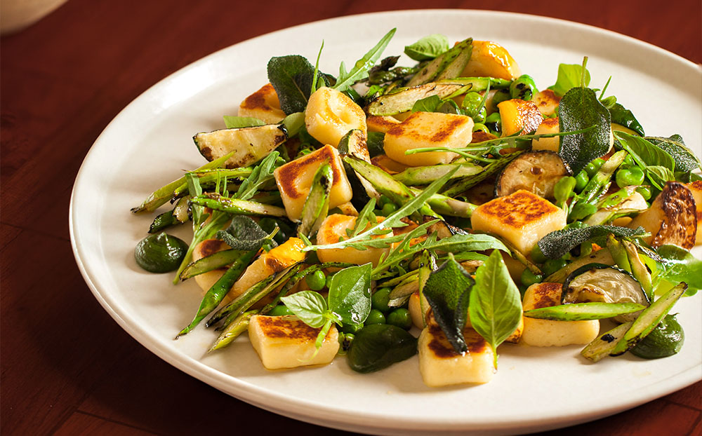 CasCades Restaurant's pan-seared gnocchi with fresh green asparagus, zucchini, peas, and arugula, garnished with sage leaves.